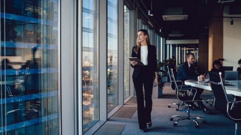 Serious female administrative manager in formal suit walking in office with digital tablet for checking office working process, confident woman business owner looking at window passing with touchpad
