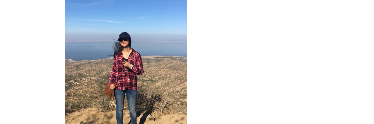 Priyanka standing in front of a scenic background of water and hills while wearing a plaid jacket.