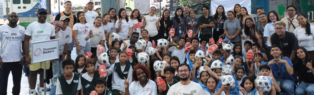 "Thomson Reuters employees volunteering with a large group of children at a community center, distributing soccer balls and sports equipment during a youth development program"